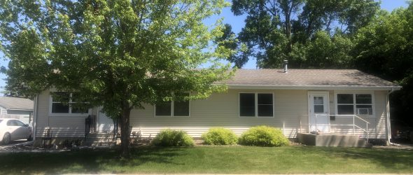 A long single-story house with light yellow siding, white trim, and a series of windows, partially obscured by green trees, with a sidewalk in the foreground under a sunny sky.